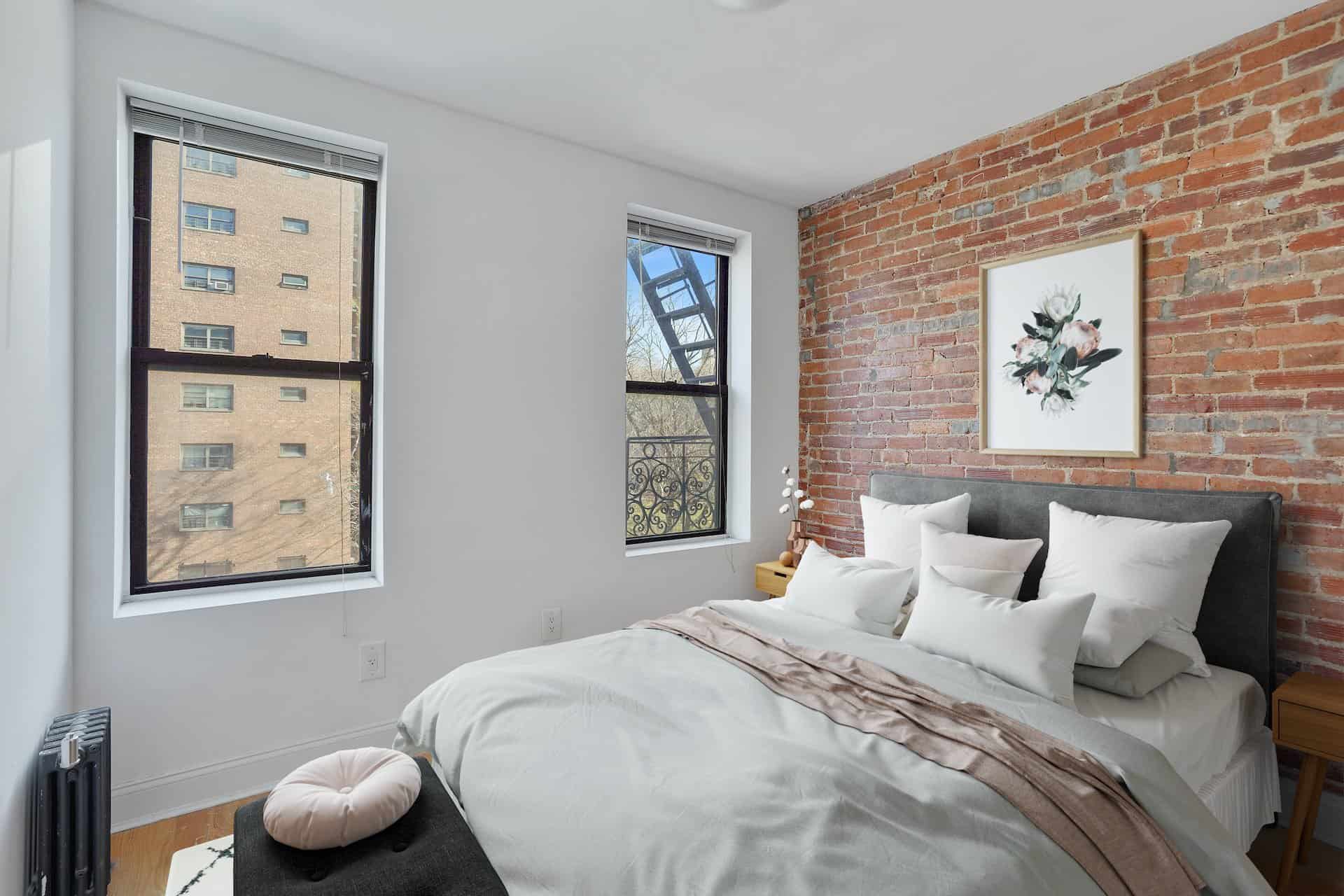 Bedroom at 556-566 West 126th Street apartments. A queen sized bed, brick accent wall, wood side tables and two windows.