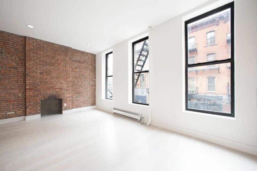 Living room at 324 Bowery Street apartments with 3 tall windows, hardwood floors, and brick accent wall with a fireplace.