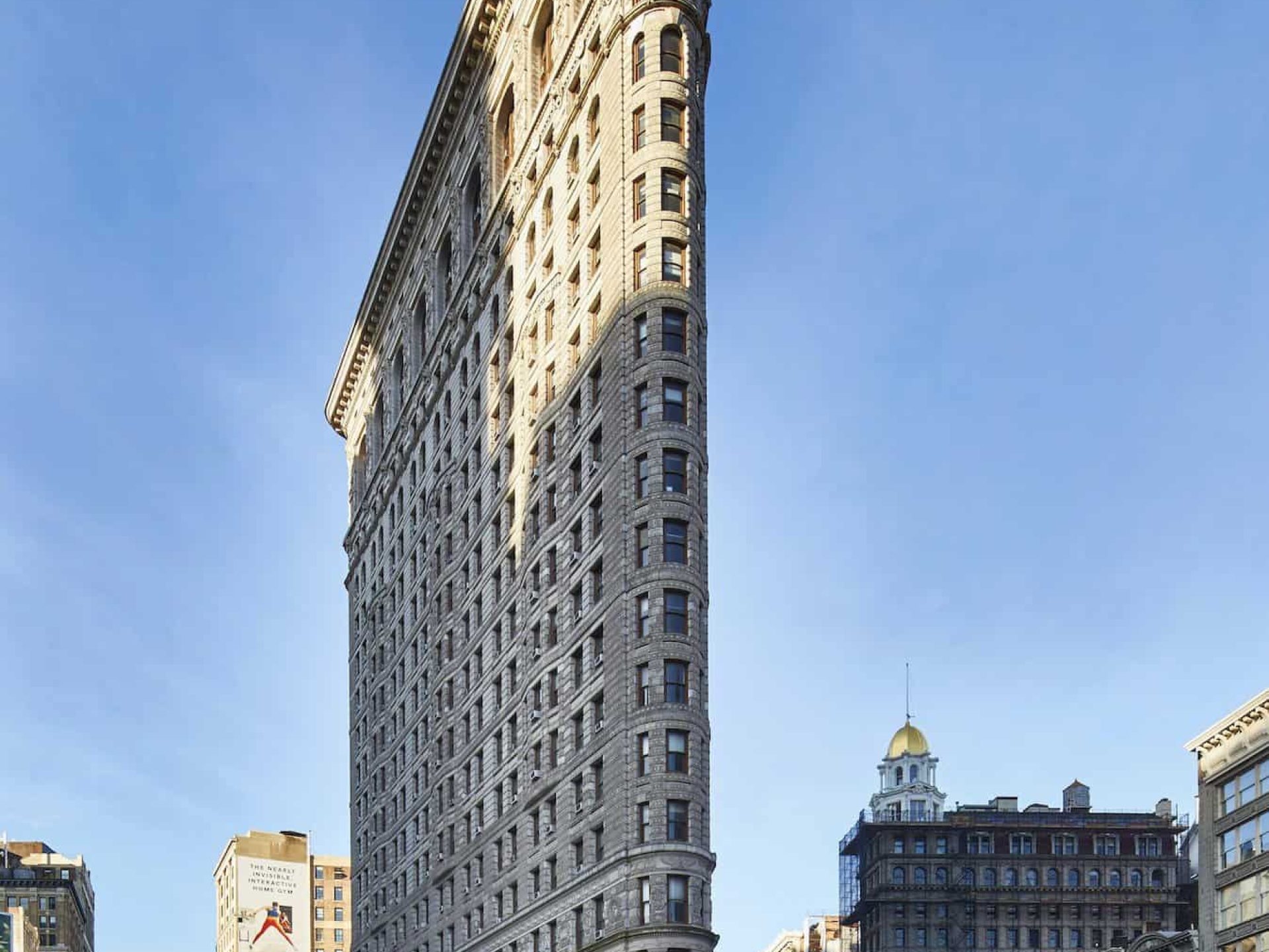 Street view of the Flatiron Building in New York. Tall skinny stone building with a green awning around the first floor.