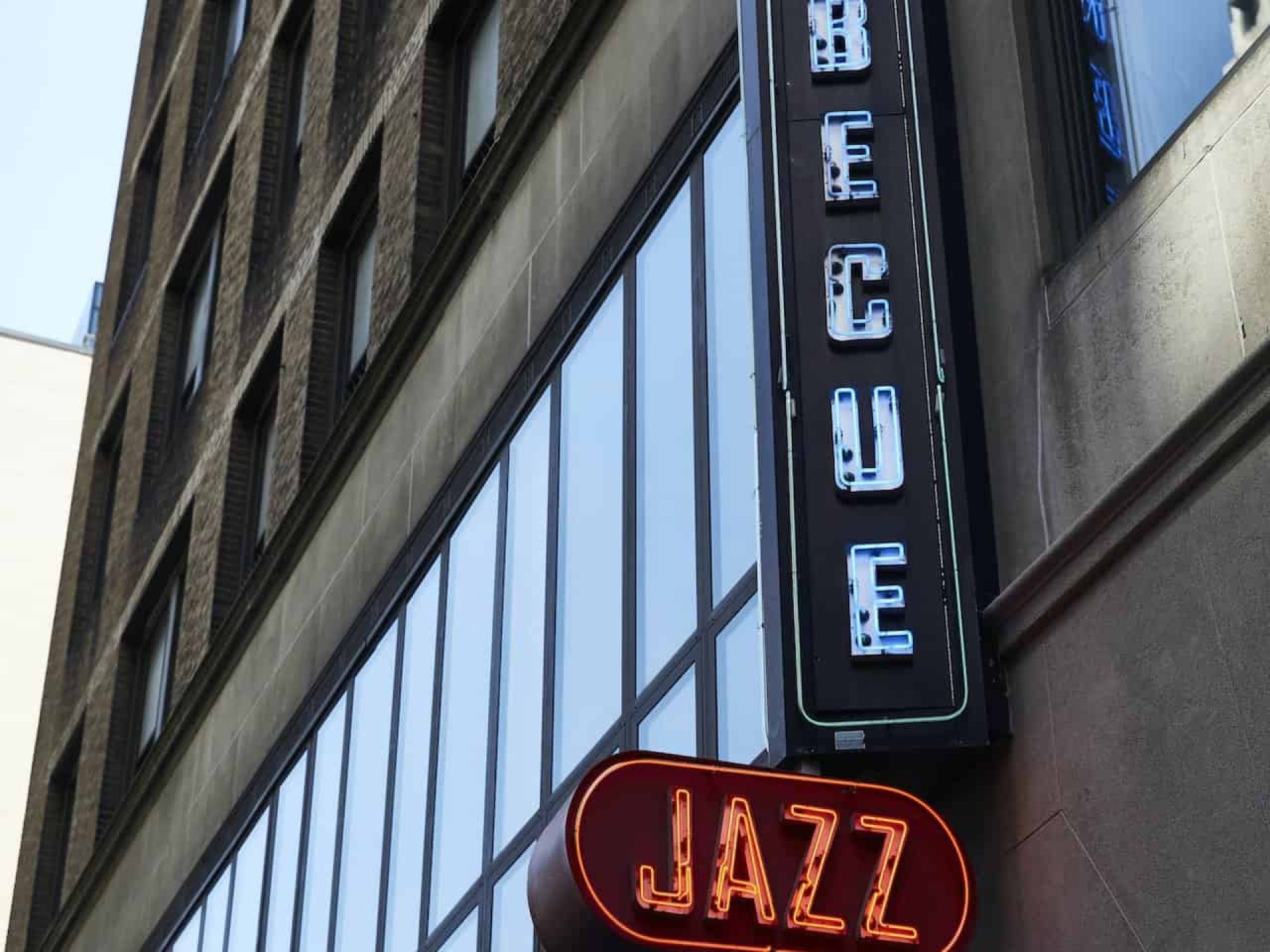 Close up of building exterior with two neon signs for "Barbecue" and "Jazz" mounted to a stone wall above building entrance.