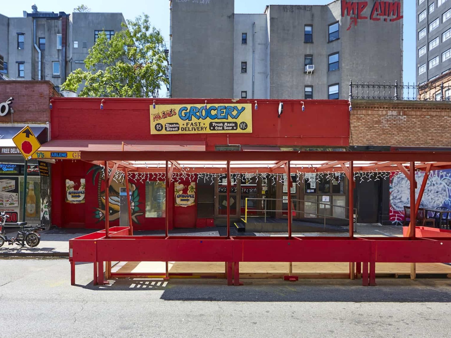 Street view of Arlene's Grocery with a ramp to entry, red painted exterior, and covered outdoor space with string lights.