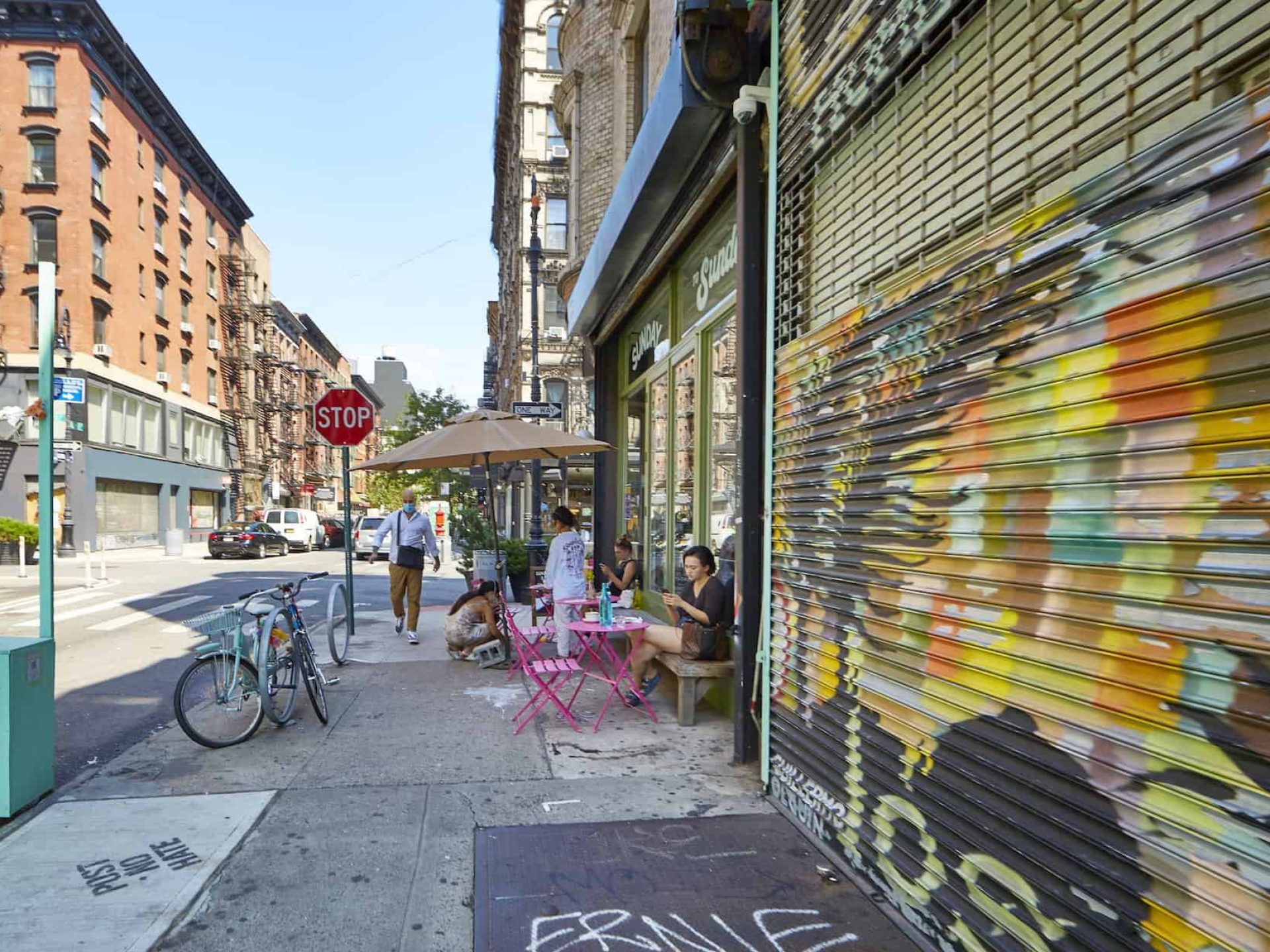 Sidewalk in New York with metal security door painted in street art and people sitting outside of a restaurant on a bench.