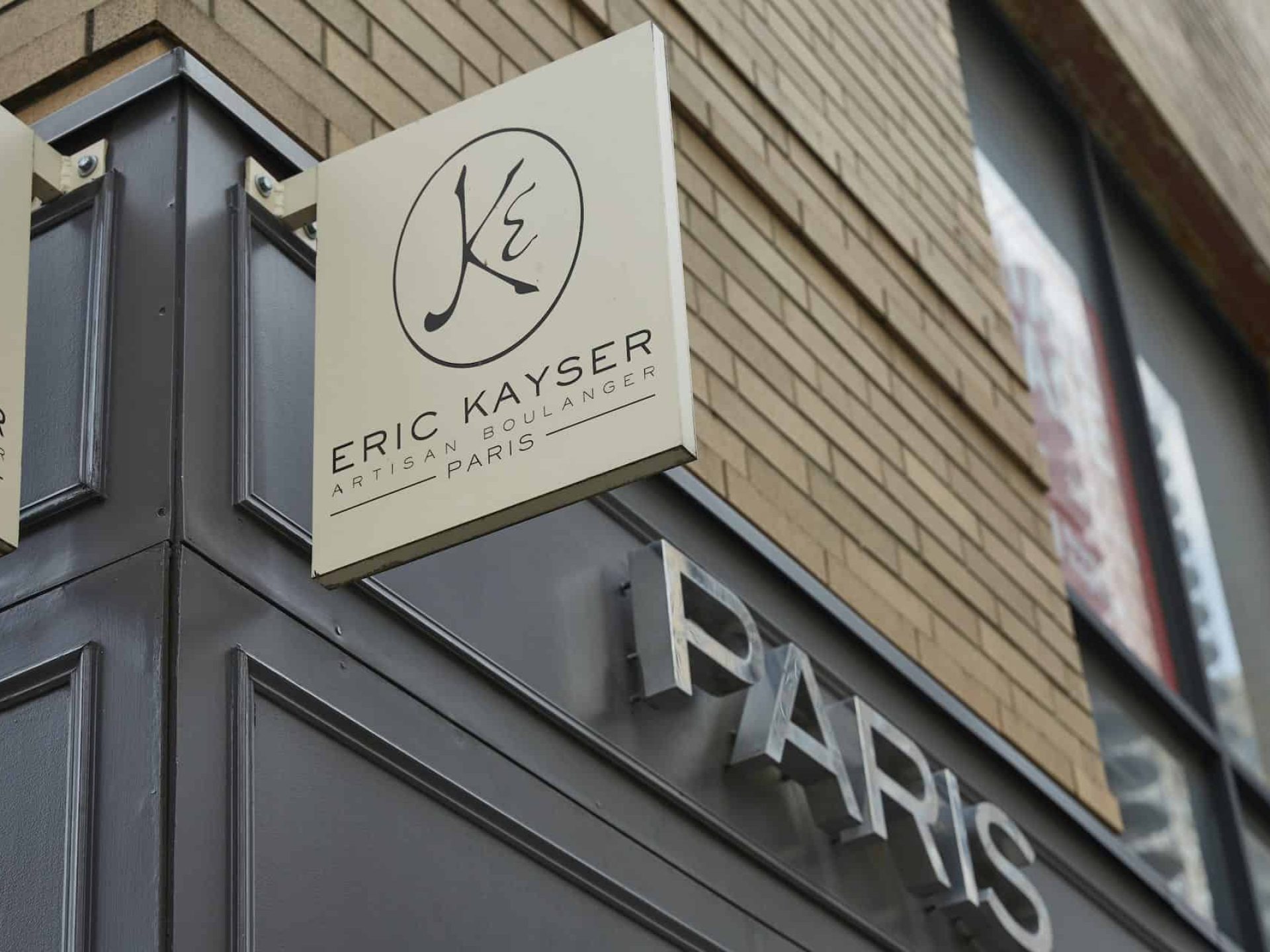 Close up of Eric Kayser Artisan Boulanger Paris business sign mounted on brick building with "Paris" sign in the background.