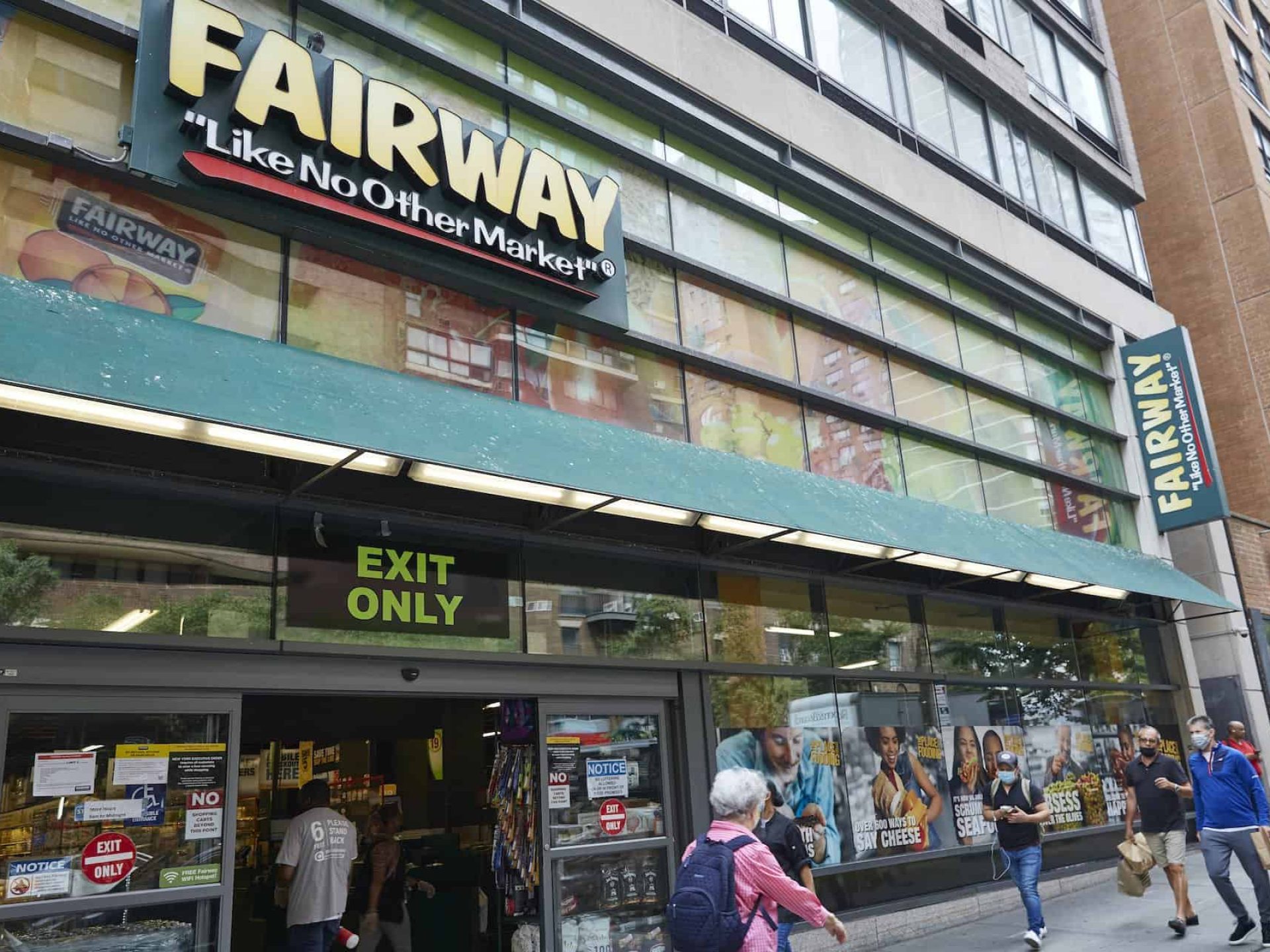 Entrance to Fairway Market with business signs, a green awning of the sidewalk, open double doors and people walking.