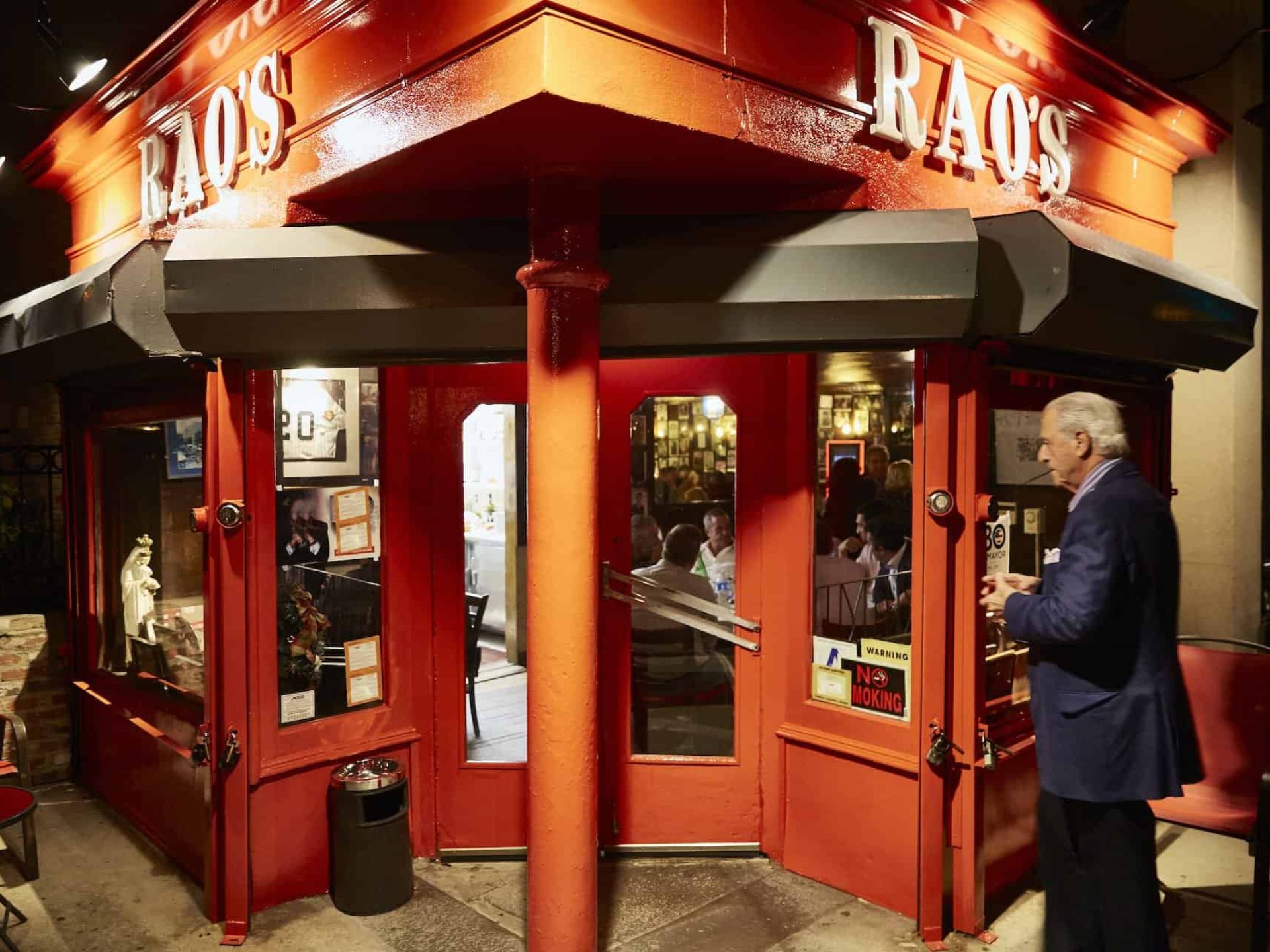Entrance to Rao's Italian restaurant in East Harlem. Red overhang and building with lit entry and a man talking to the door.