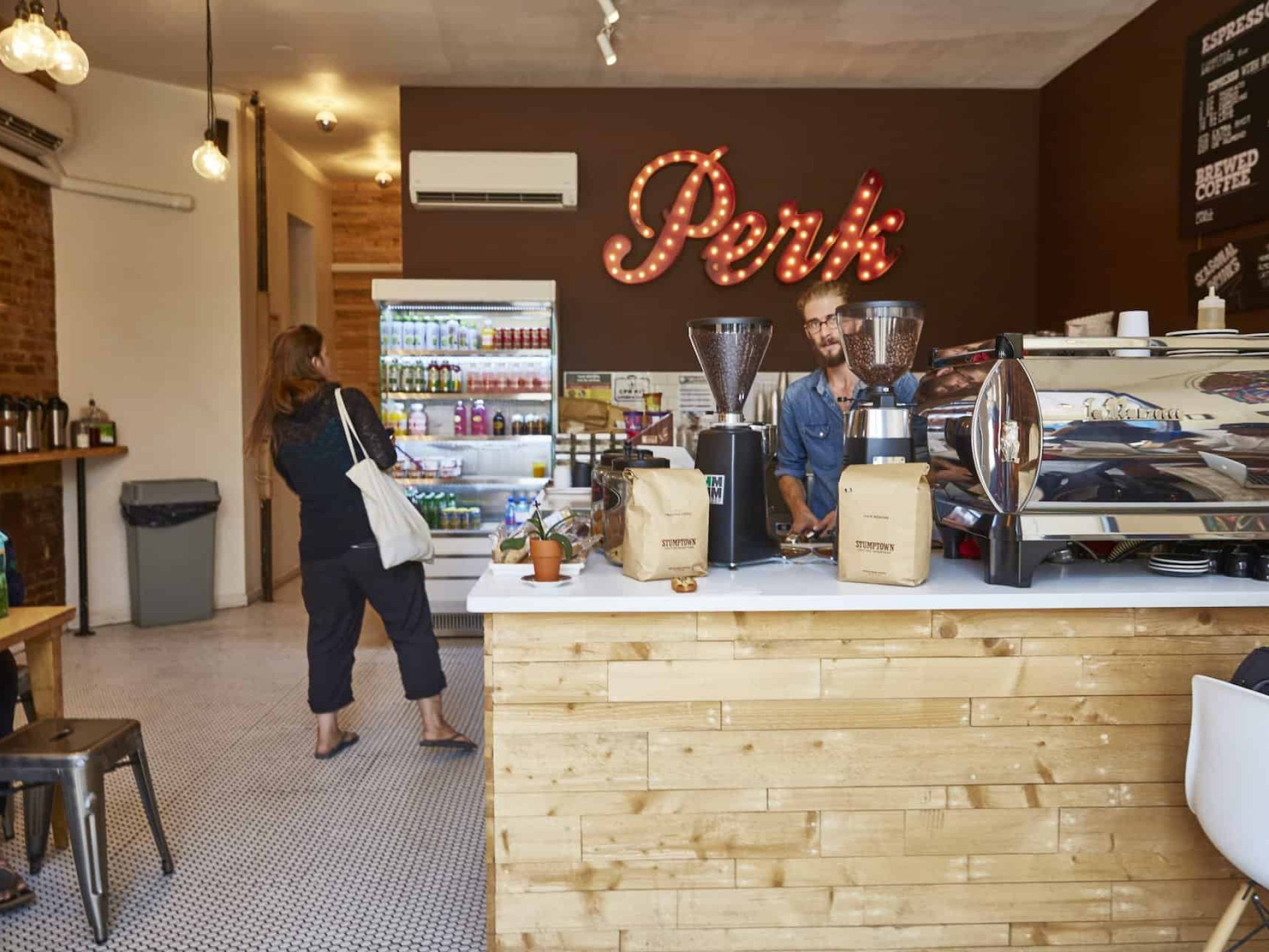 Interior of "Perk Coffee" shop with business sign on the back wall and a white L-shape counter with coffee and grinders.