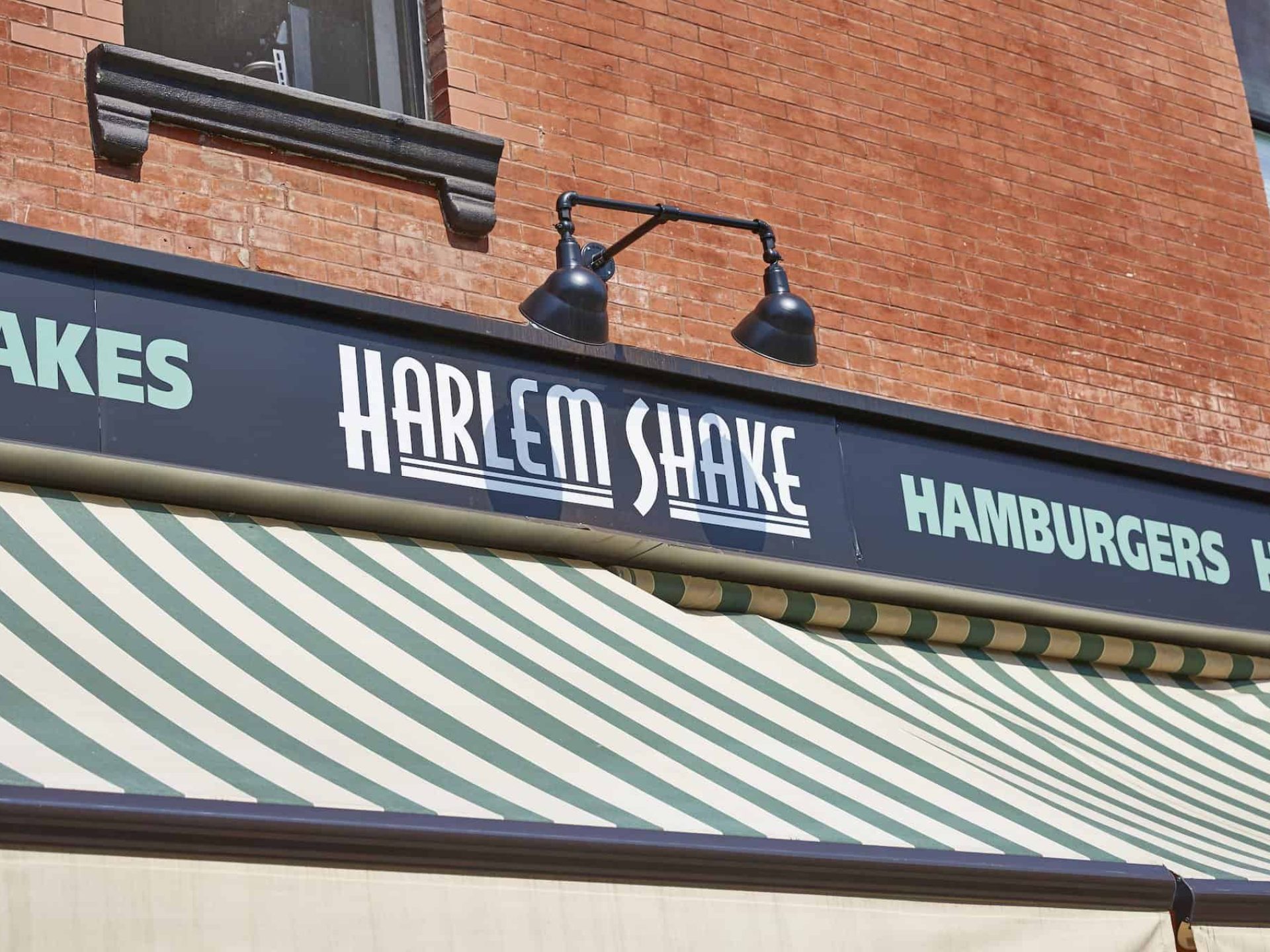 Close up of "Harlem Shake" restaurant with mounted lights above business sign and a striped awning below the sign.