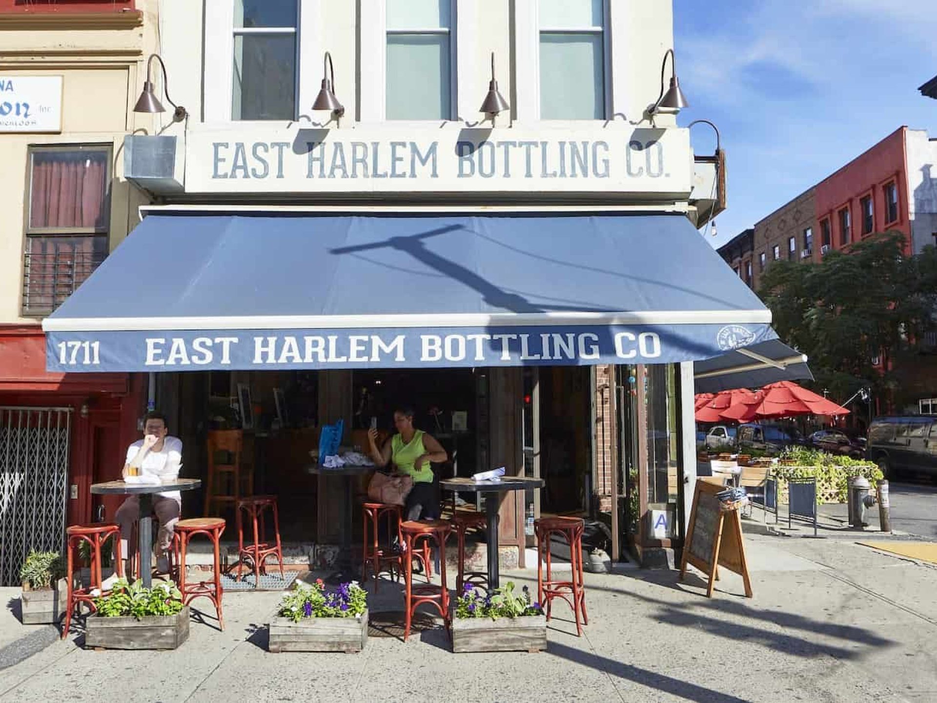 Exterior of East Harlem Bottling Co. in New York. Floor corner unit with a blue canopy, outdoor seating area and open doors.
