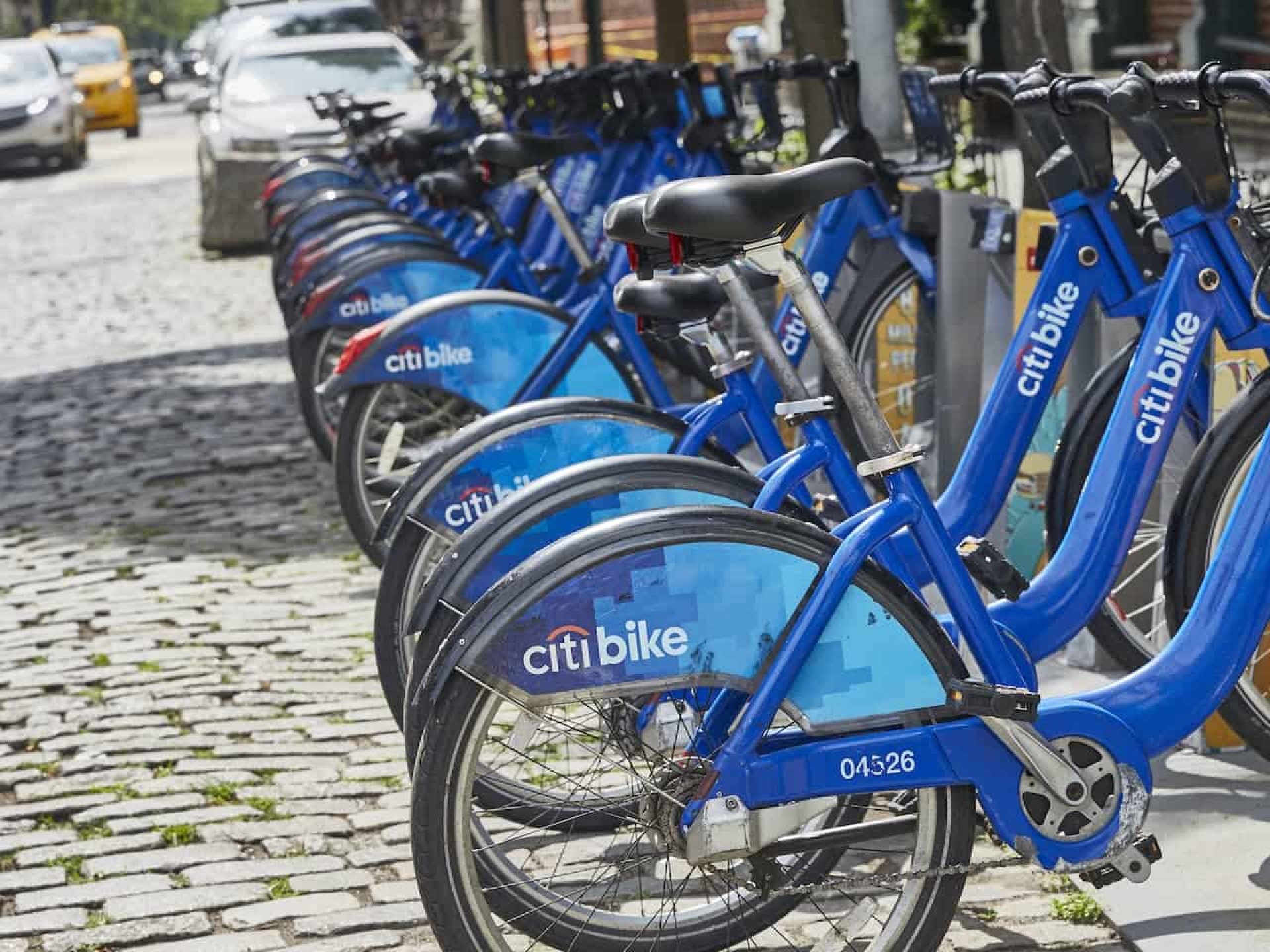 Blue Citi bikes lined up in bike racks along a brick street in the West Village neighborhood with cars in the background.