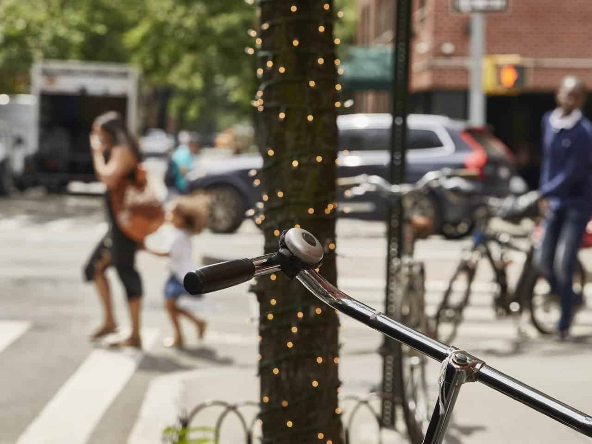 A tree planted in a sidewalk flower bed wrapped in white string lights with a bike handle in the foreground.