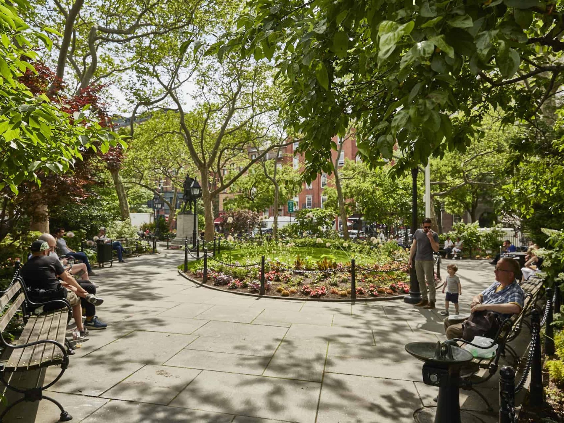Paved circular opening in a New York City park with benches bordering the path and a raised flower bed in the middle.