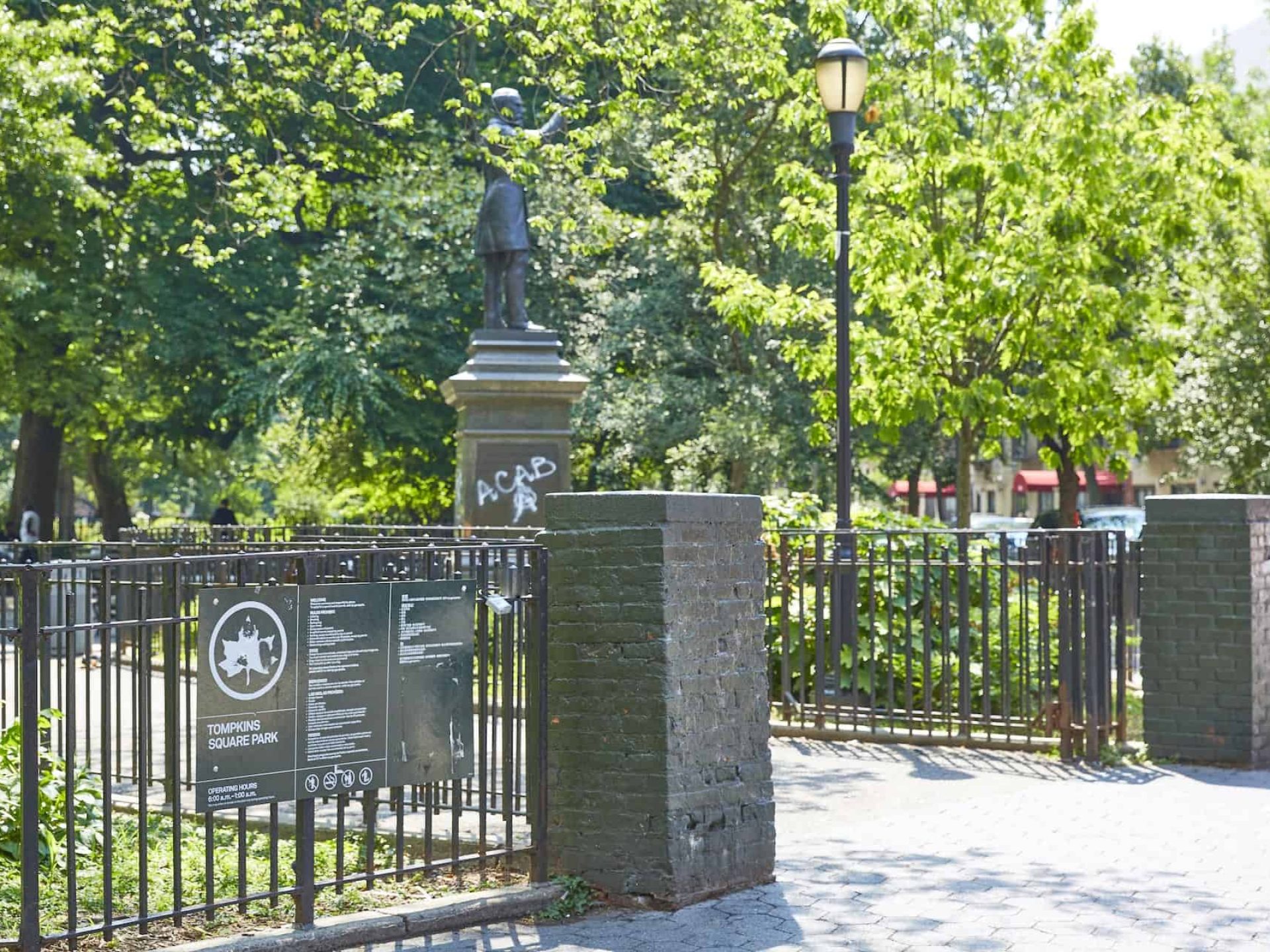 Daytime picture of entrance to Tompkins Square Park. Green iron fences with park sign attached and statue in the background.