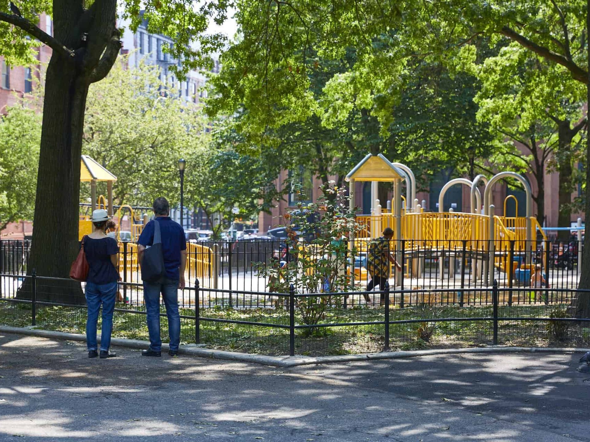 Picture of a children's playground in a city park. Two parents looking at the playground from a park path between trees.