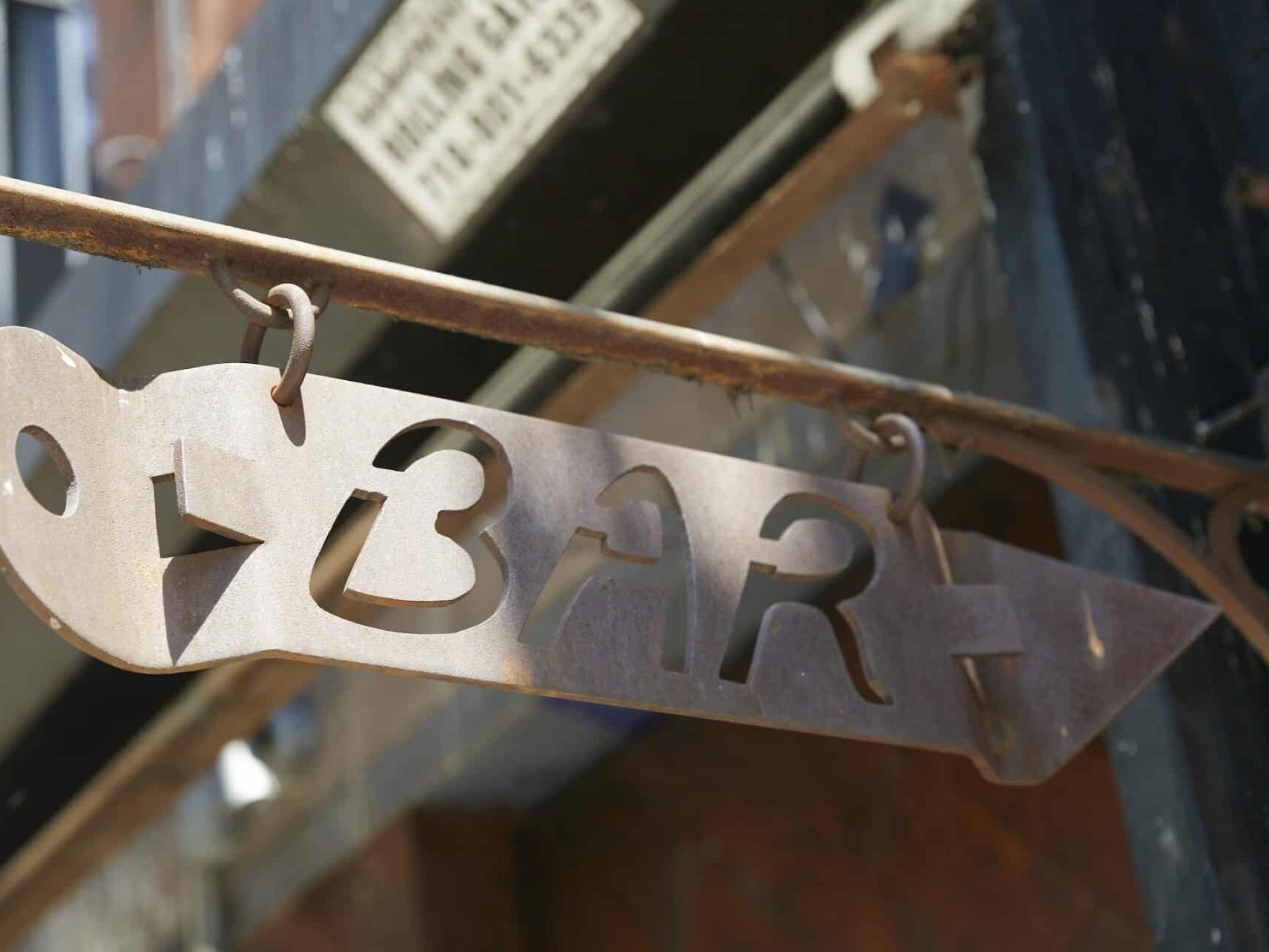 Close up of a sign hanging from a building. Brass sign in the shape of old bottle opener with "Bar" engraved in the center.