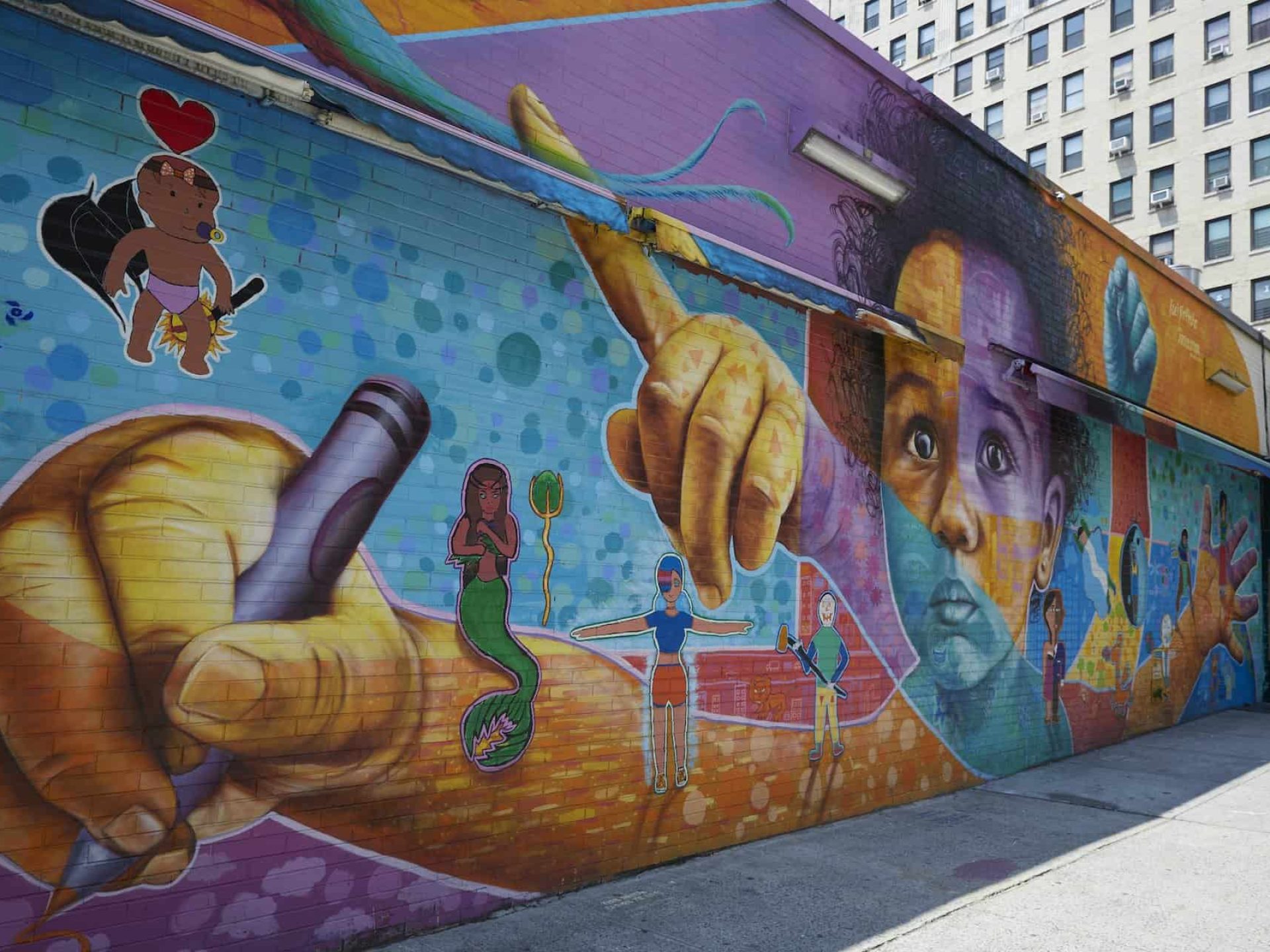 A street mural painted on the side of a brick building. Picture of a babies face, children's hands and other characters.