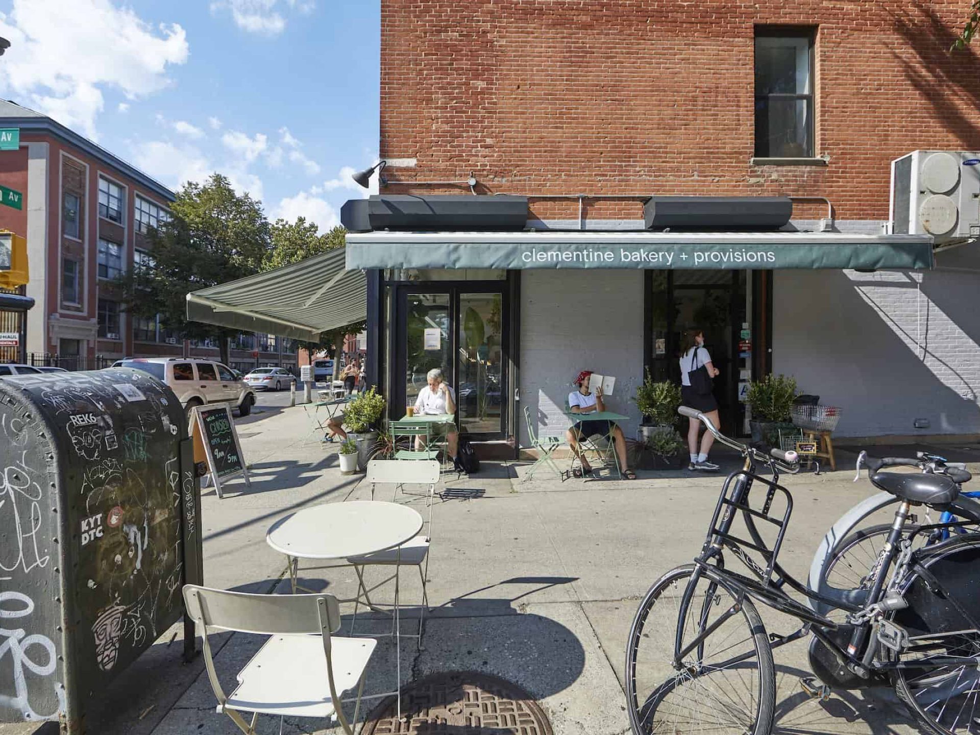 Sidewalk view of the Clementine Bakery in Clinton Hill. White painted brick wall with green awning covering outdoor seating.