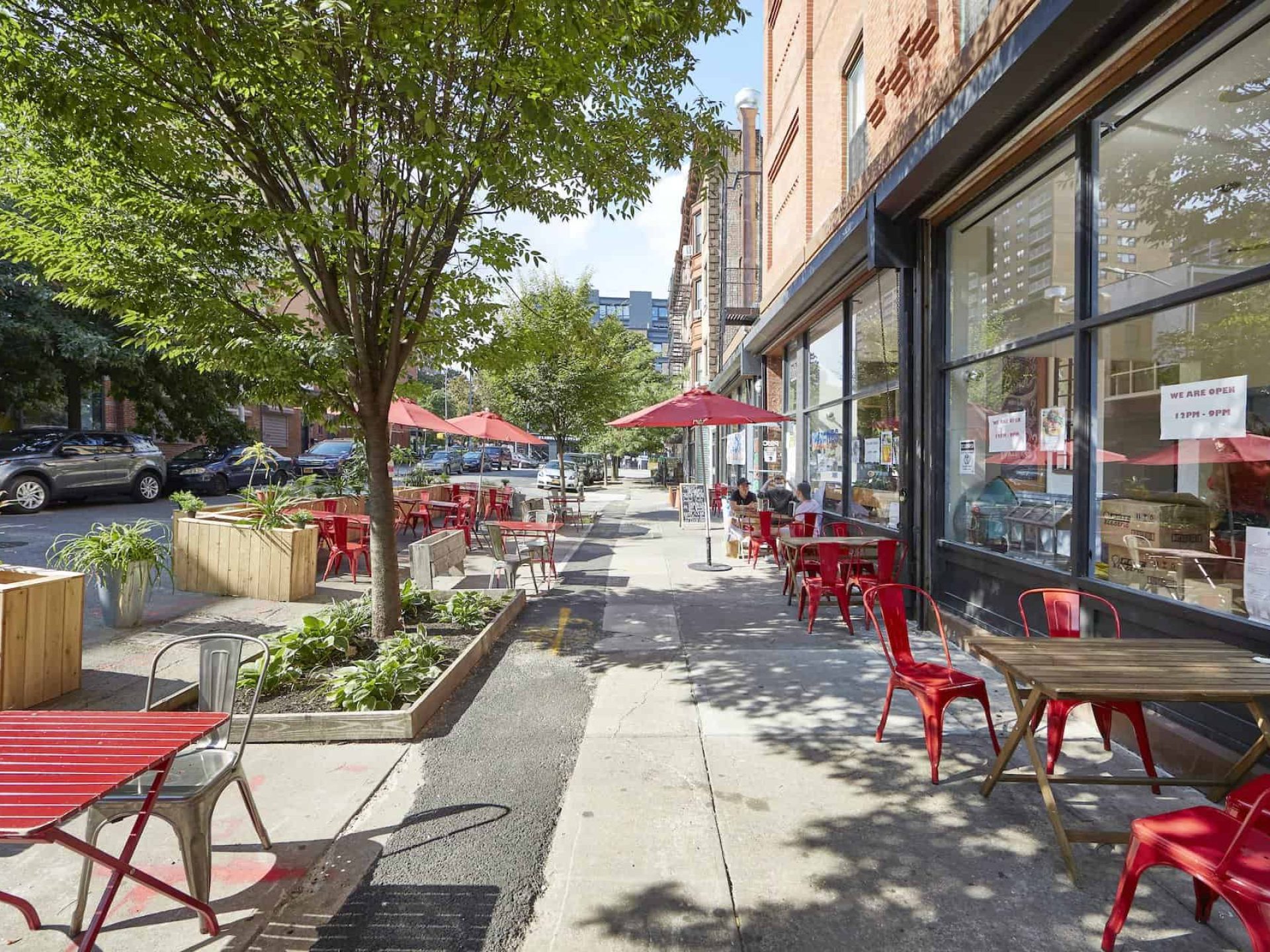 View of sidewalk lined with wood square tables and red chairs in the Clinton Hill neighborhood outside of a restaurant.