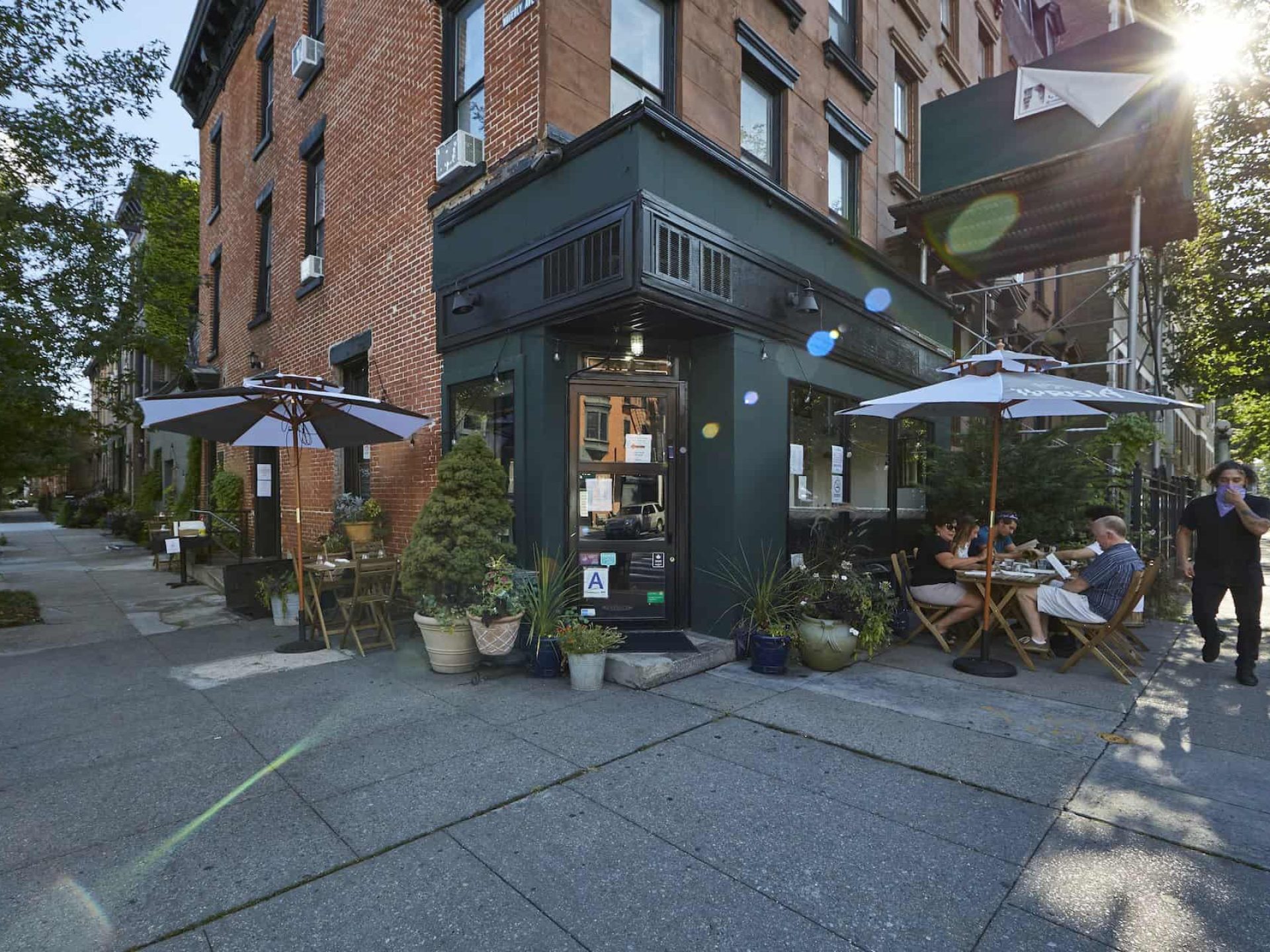 Restaurant entrance at the corner of a brick building with a green exterior and outdoor seating area with umbrellas.