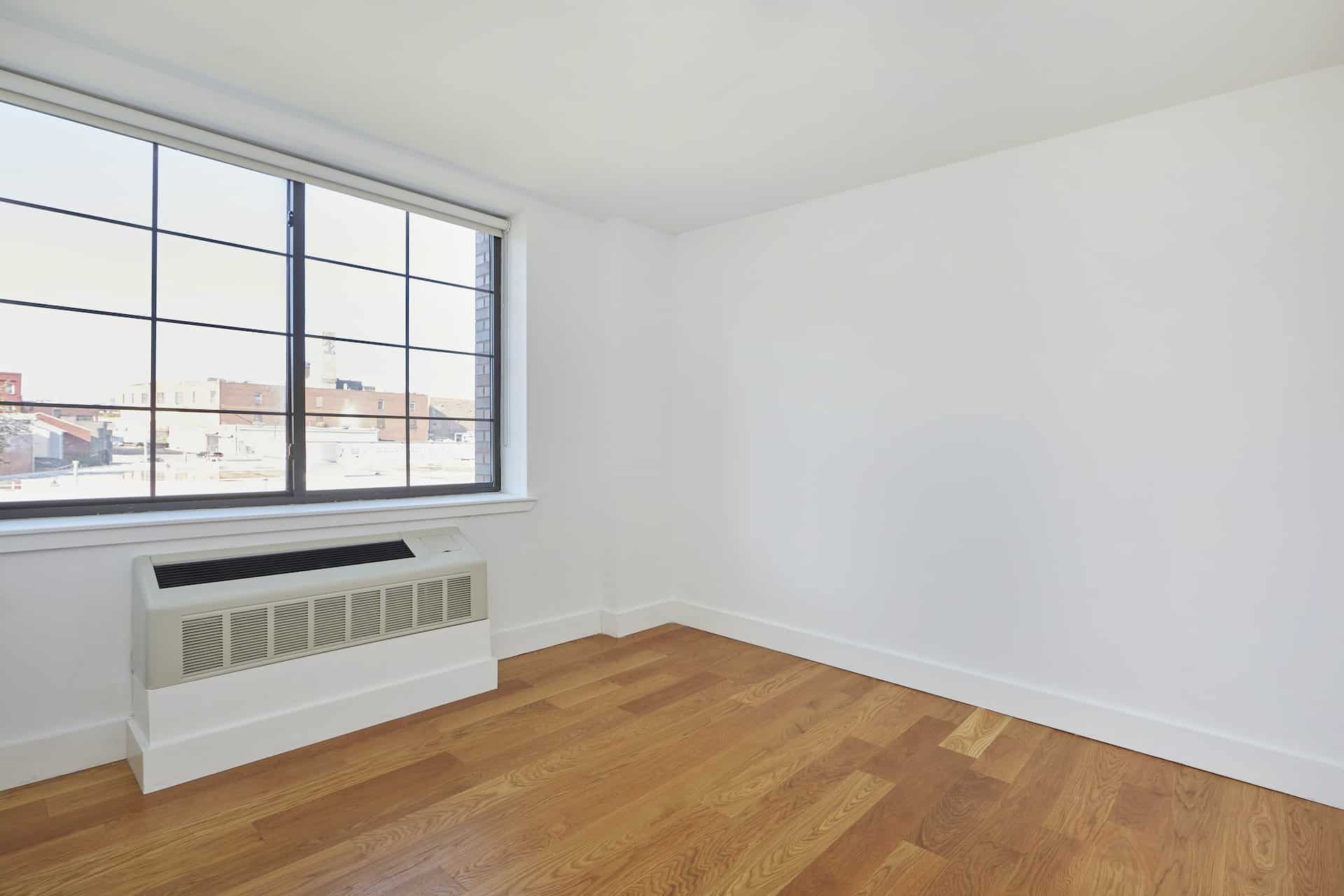 Living room at 83 Bushwick Place apartments in Brooklyn with hardwood floors, floor mounted a/c unit and large windows.