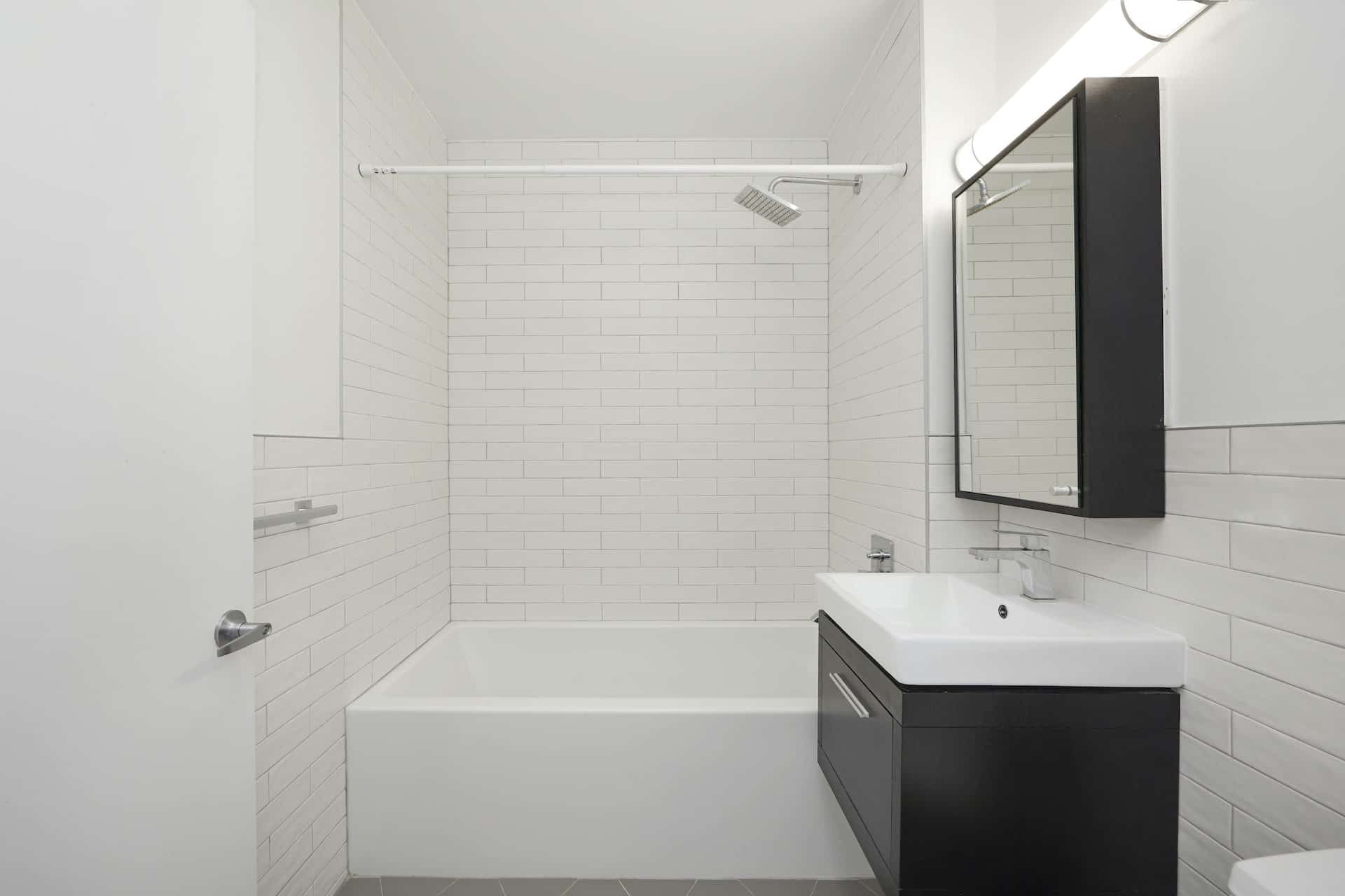 Bathroom at 83 Bushwick Place apartments with a single vanity, square mirror, tile walls, and a soaking tub with shower.