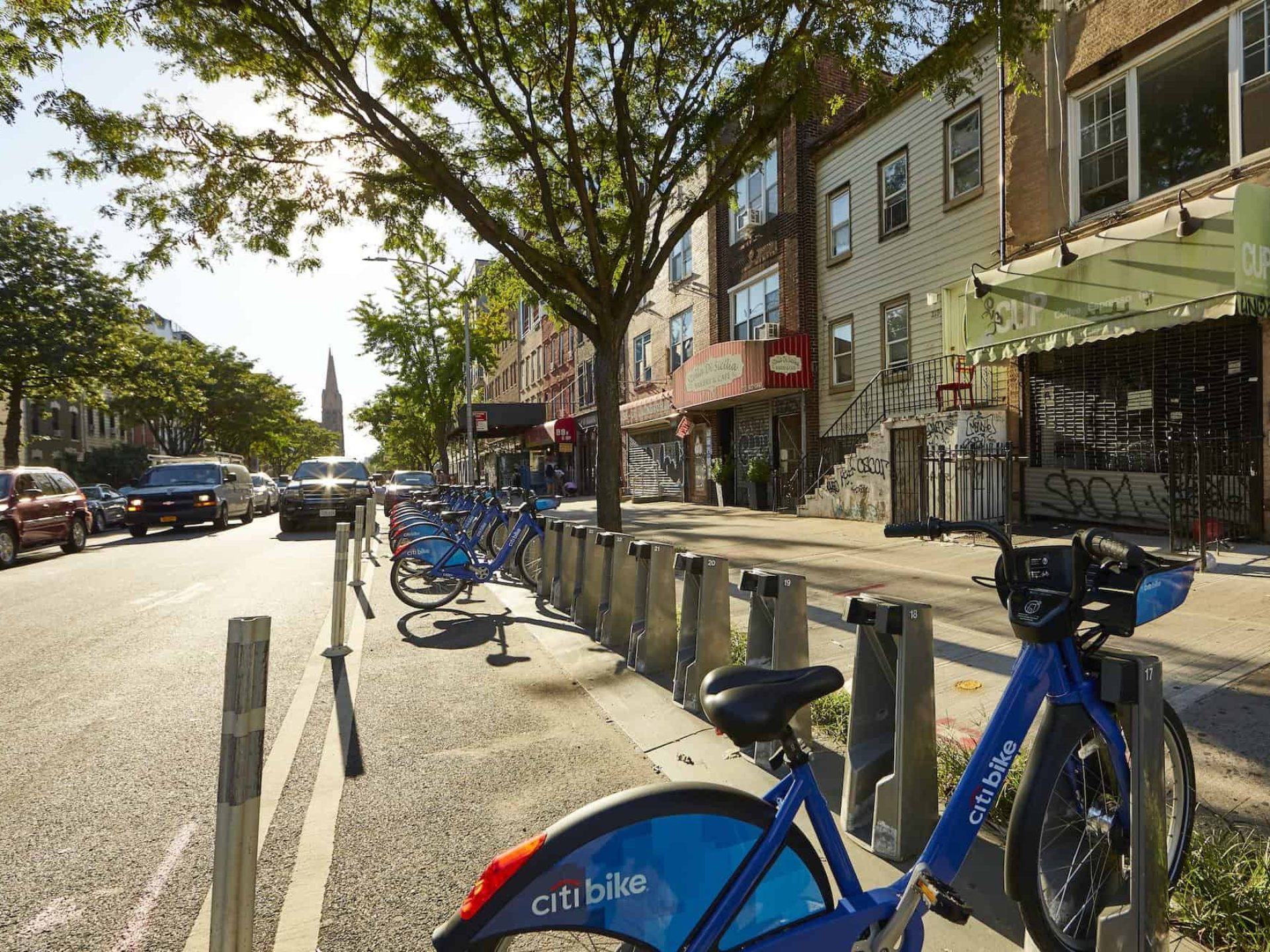 Blue Citi bikes lined up in bike racks along a street in the East Williamsburg neighborhood with cars in the background.