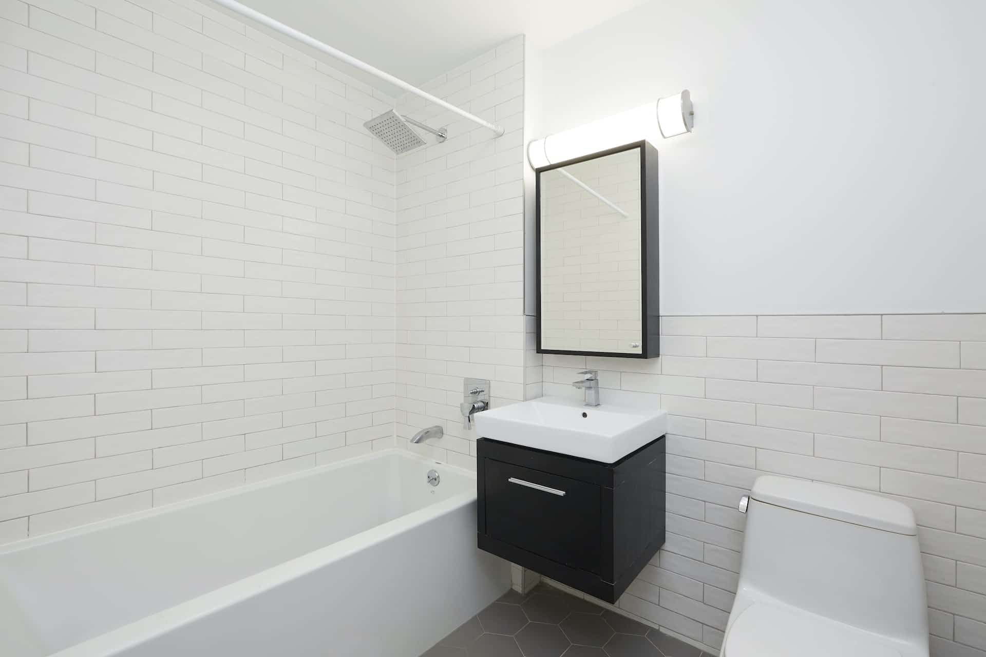 Bathroom at 83 Bushwick Place apartments with single vanity, a mirror, tile walls & floor, and a soaking tub with shower.