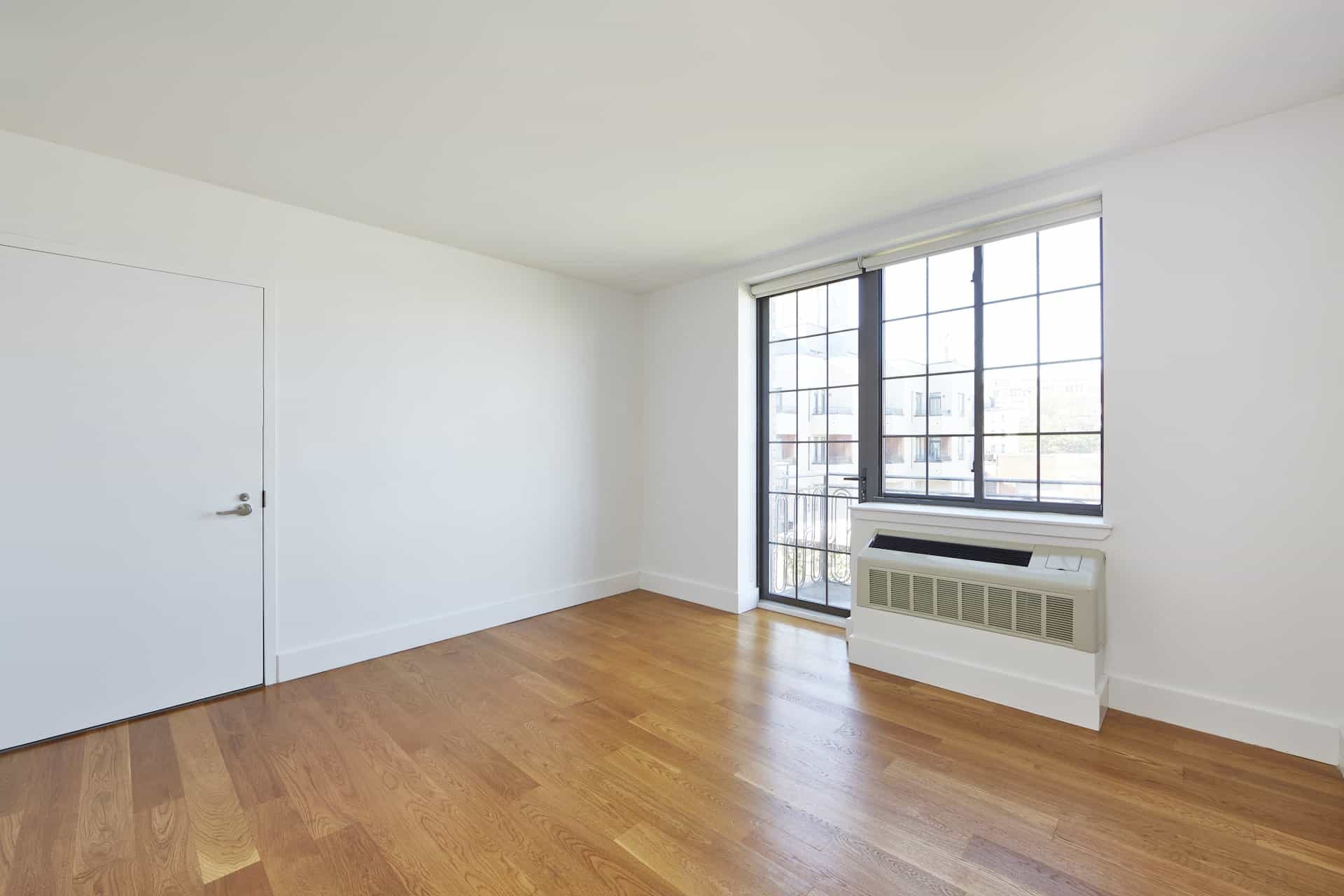 Living room at 83 Bushwick Place apartments in Brooklyn with hardwood floors, a/c unit, large windows and door out to patio.